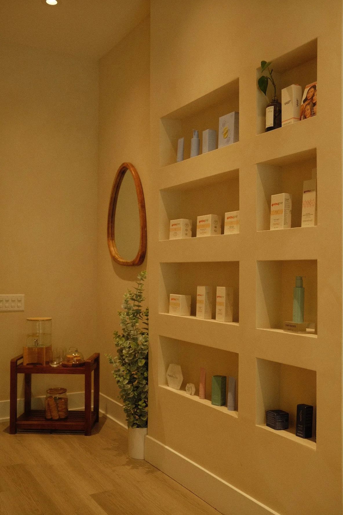 A beige wall with built-in shelves displaying skincare and beauty products, a wooden mirror, a small table with bottles and jars, and a tall potted plant.