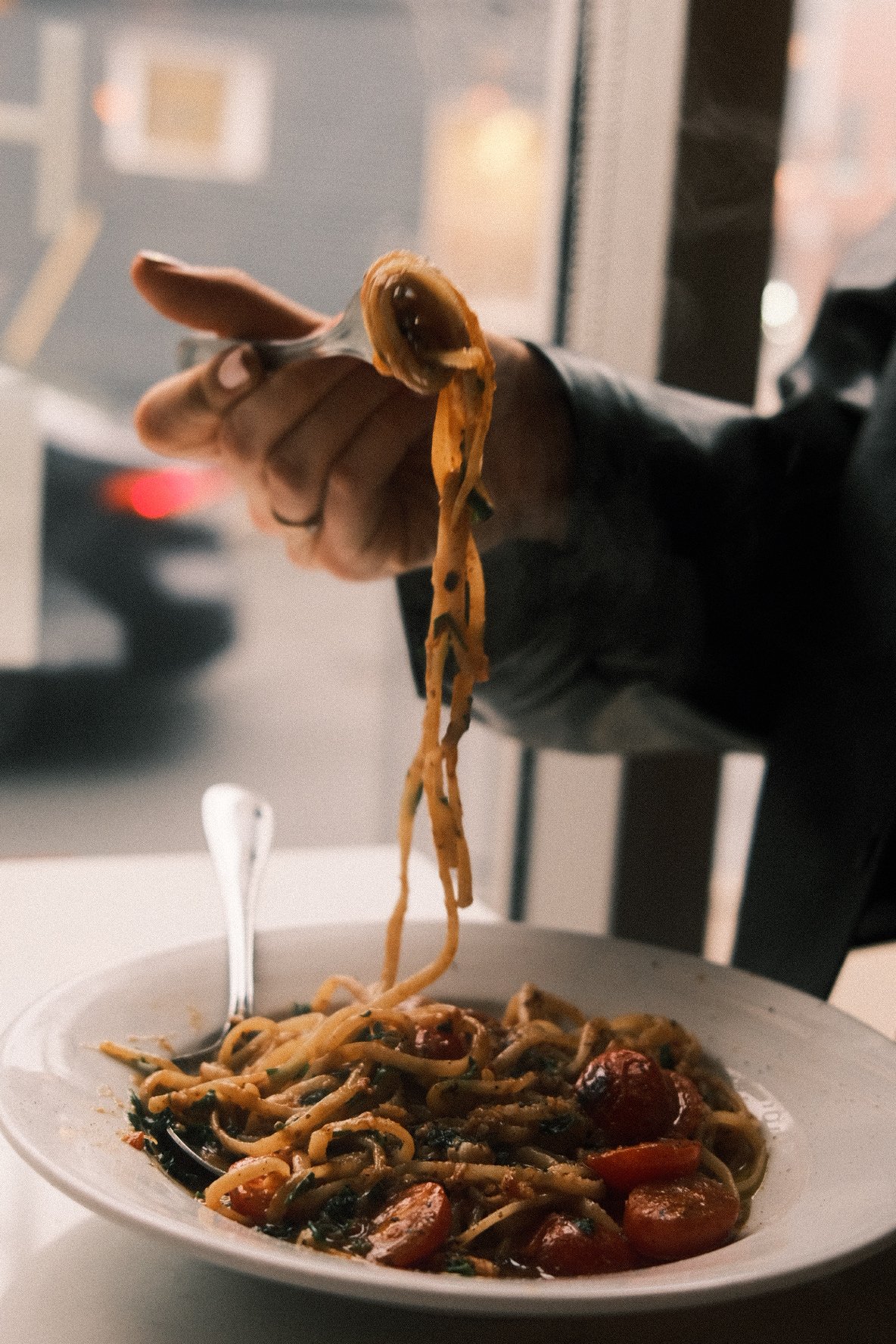 A person holding a fork with spaghetti and tomato sauce above a plate of spaghetti with cherry tomatoes and green herbs.