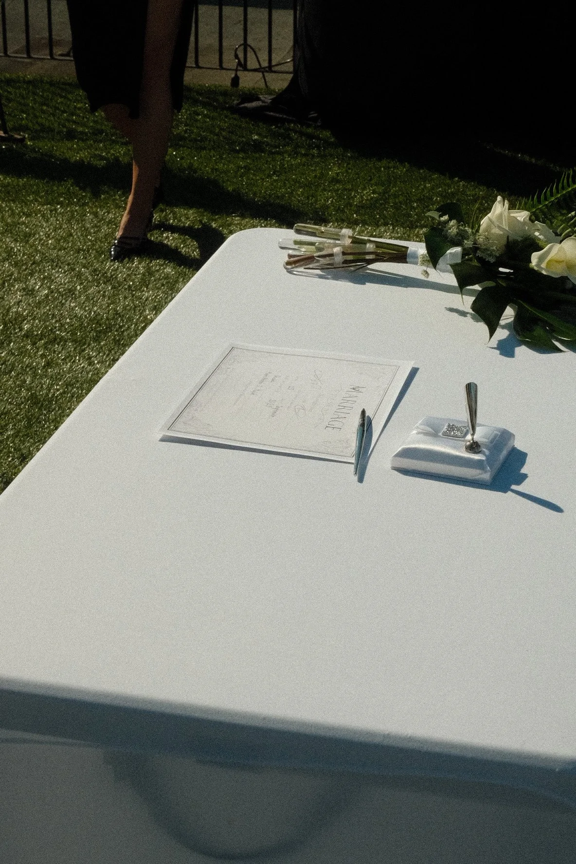 A memorial table with a framed certificate, flowers, and small ceremonial items, set outdoors on a white surface, with a person walking nearby on a grassy area.