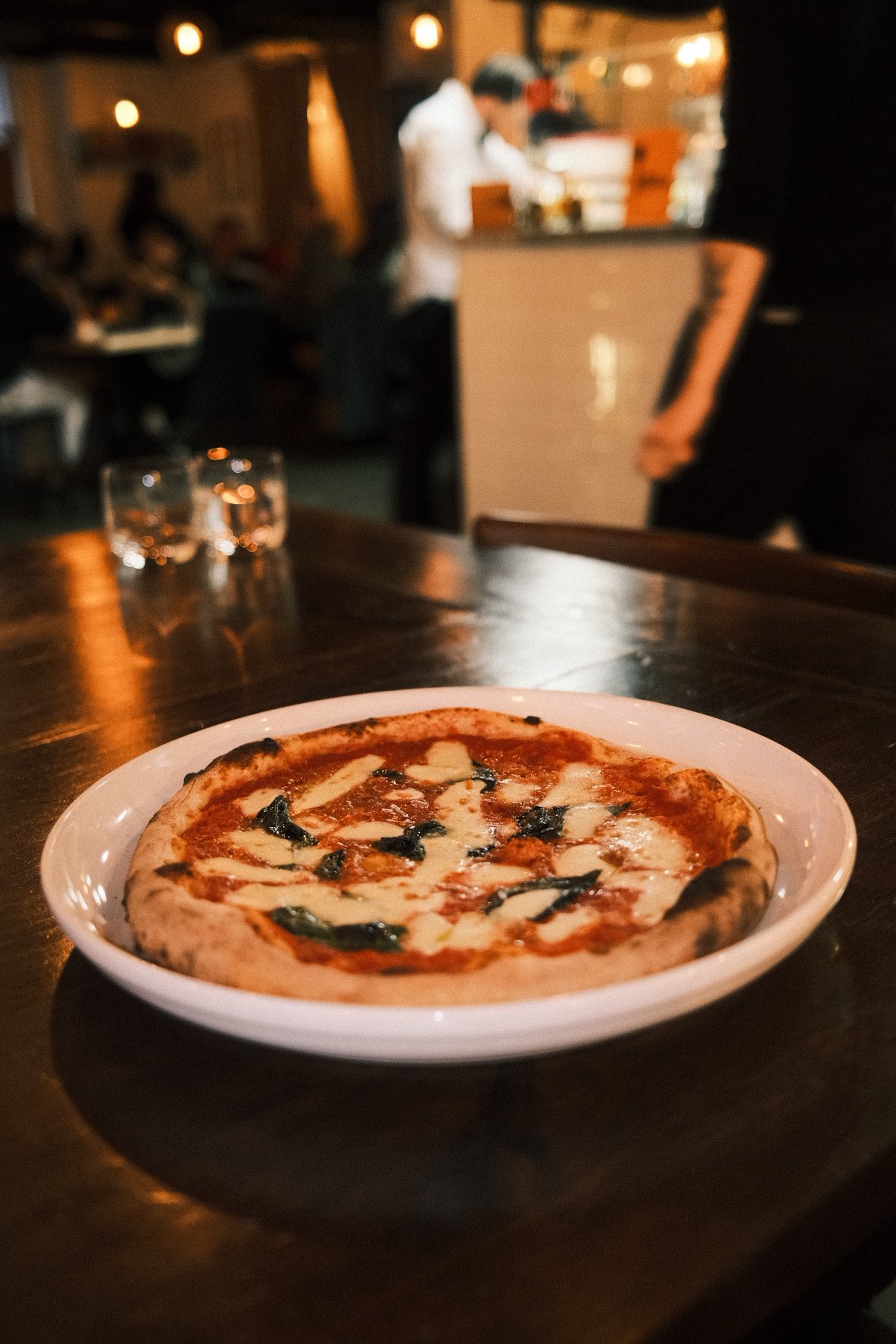 A pepperoni and cheese pizza on a white plate on a dark wooden table, with a blurred background of a restaurant and a person working behind the counter.