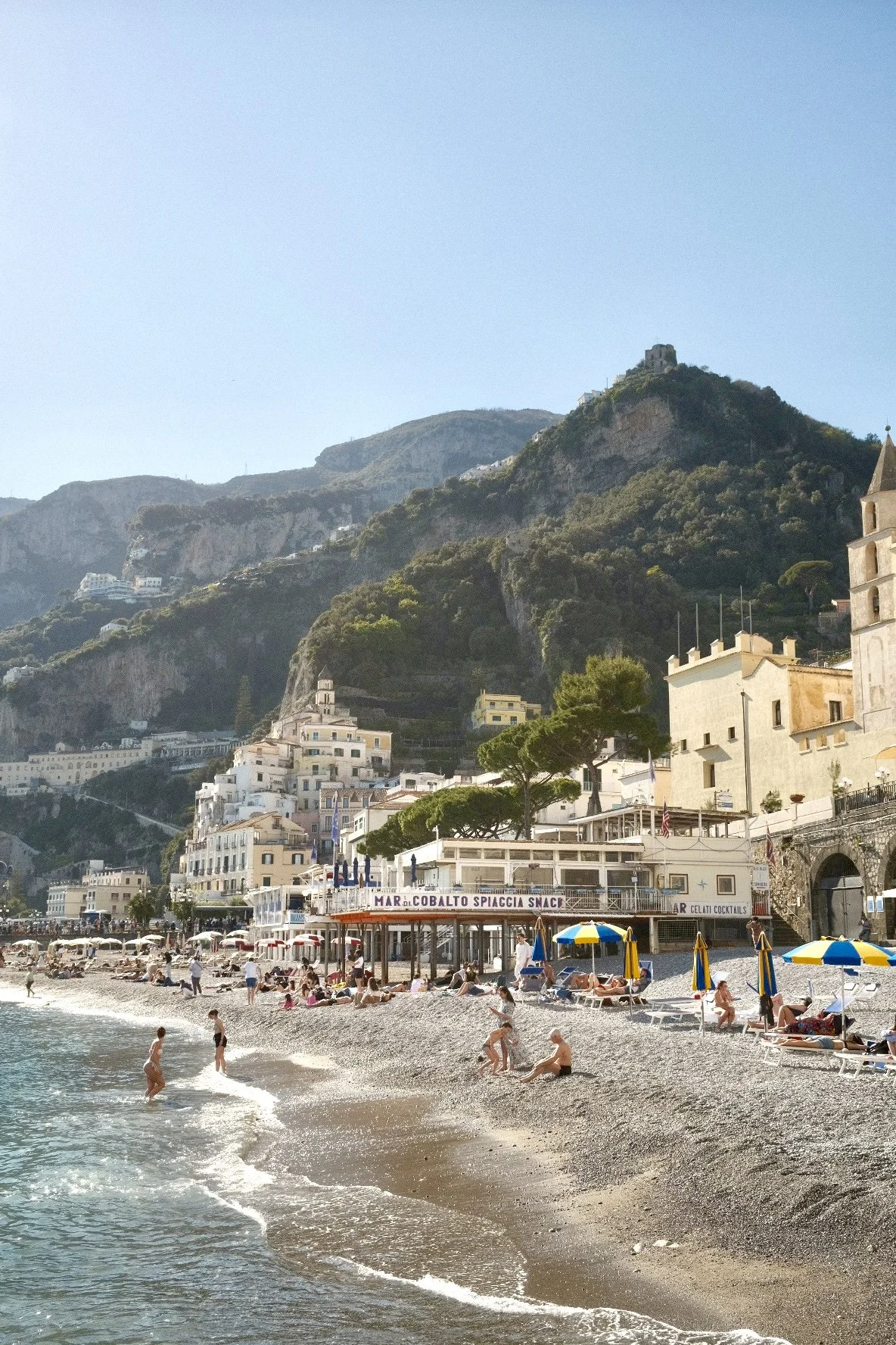 Beach with people swimming and relaxing under colorful umbrellas, a building with a sign that reads "Cobalto Spiaggia Snack," and hillside buildings with greenery and rocky terrain in the background.
