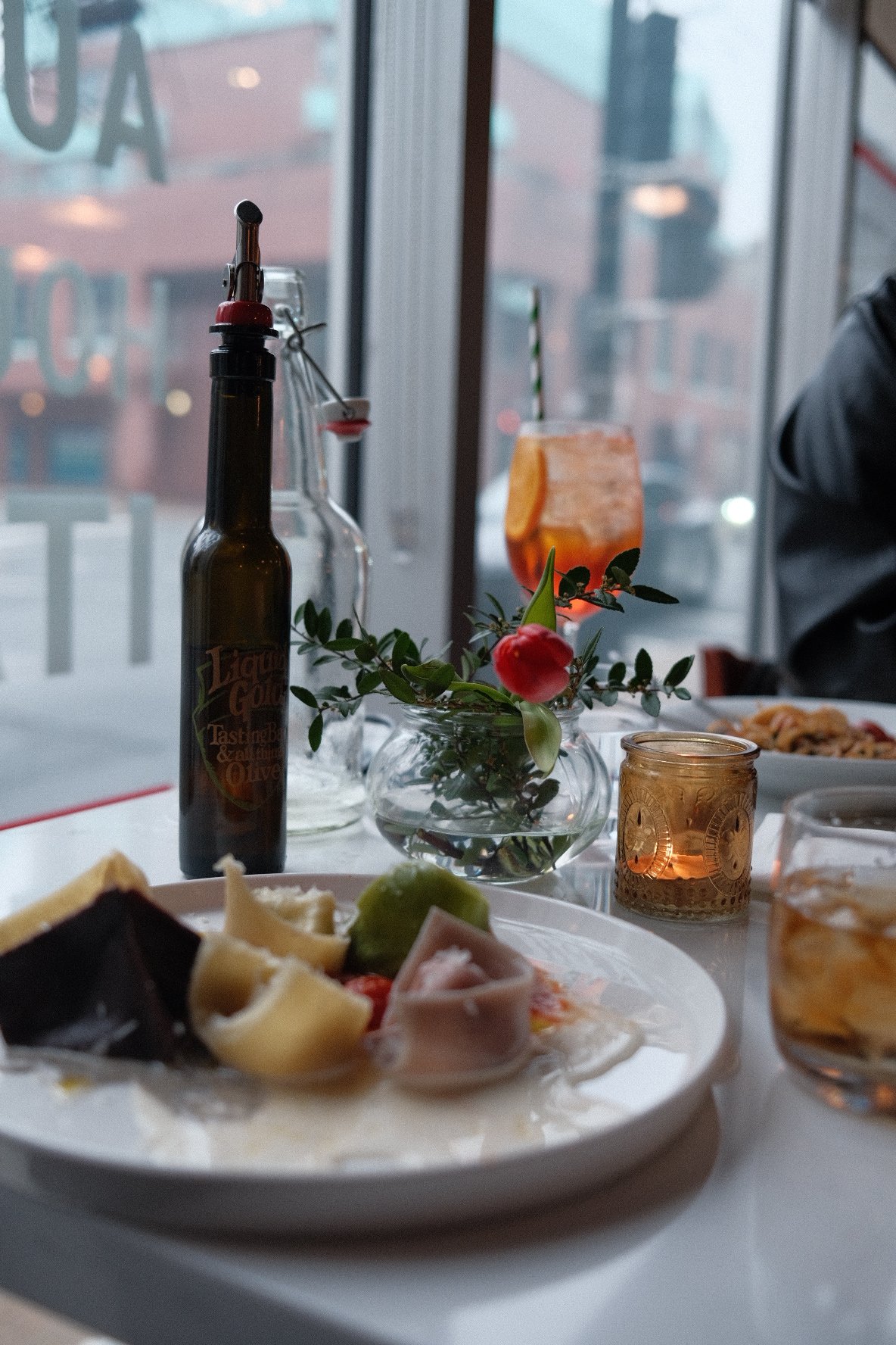 A table with a plate of assorted cheeses and meats, a bottle of hot sauce, a small glass jar, a clear vase with a red flower and greenery, a candle holder, and a drink in the background outside a window.