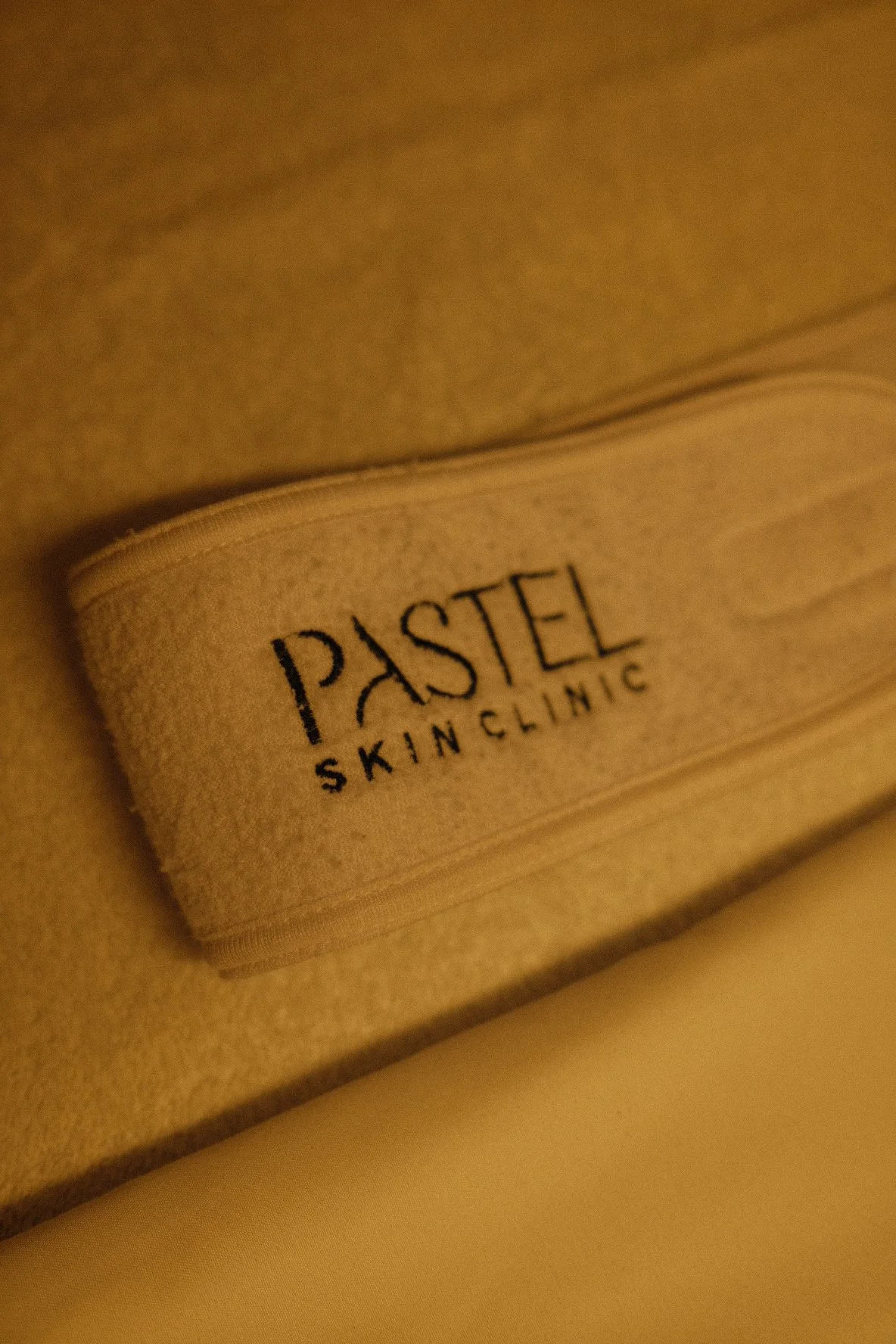 Close-up of a beige fabric headband with the words "PASTEL SKIN CLINIC" embroidered in black.