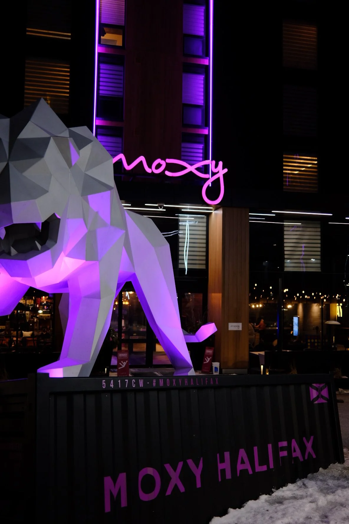 Night view of Moxy Halifax hotel with a large illuminated white geometric lion sculpture in front, purple neon signage, and the building with purple and yellow lighting in the windows.