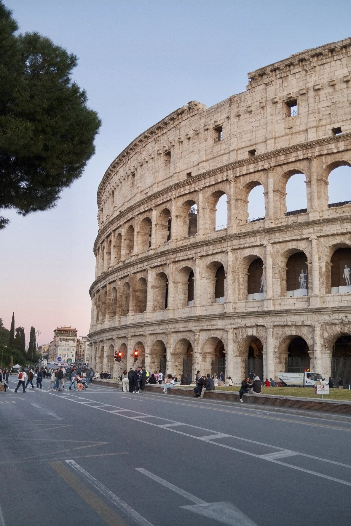 The image shows the Colosseum in Rome at dusk with many people walking and sitting outside, and a clear sky in the background.