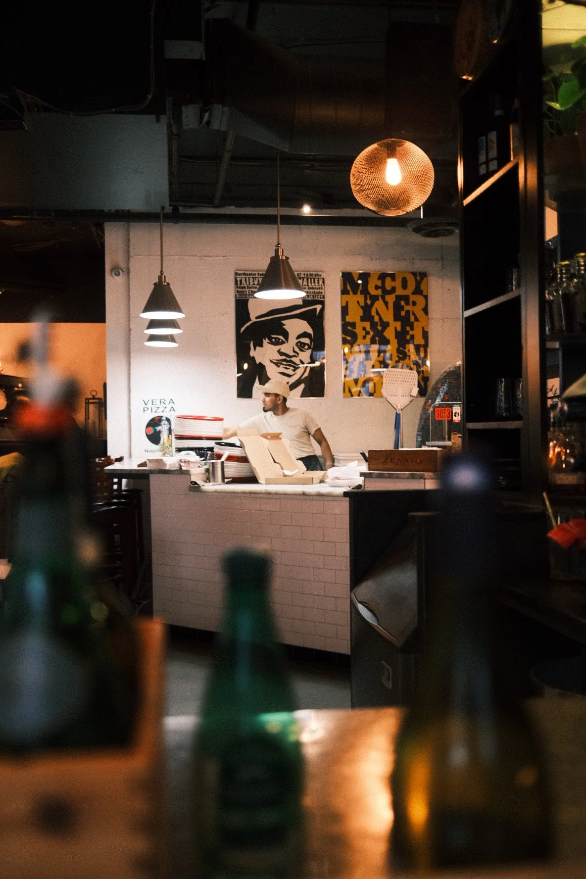 A man working behind the counter at a pizzeria, with artwork on the wall and pendant lights hanging above.