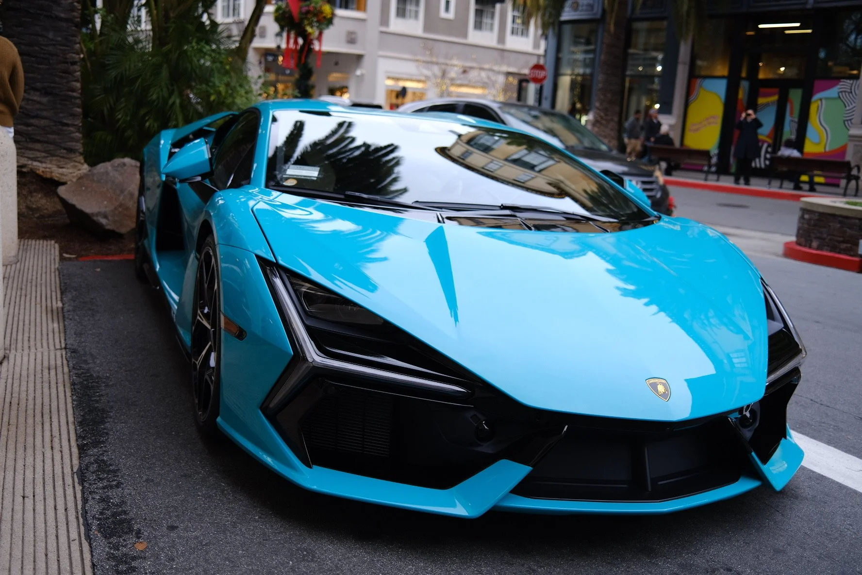 A bright blue Lamborghini sports car parked on a city street, reflecting surrounding buildings and trees on its shiny surface.