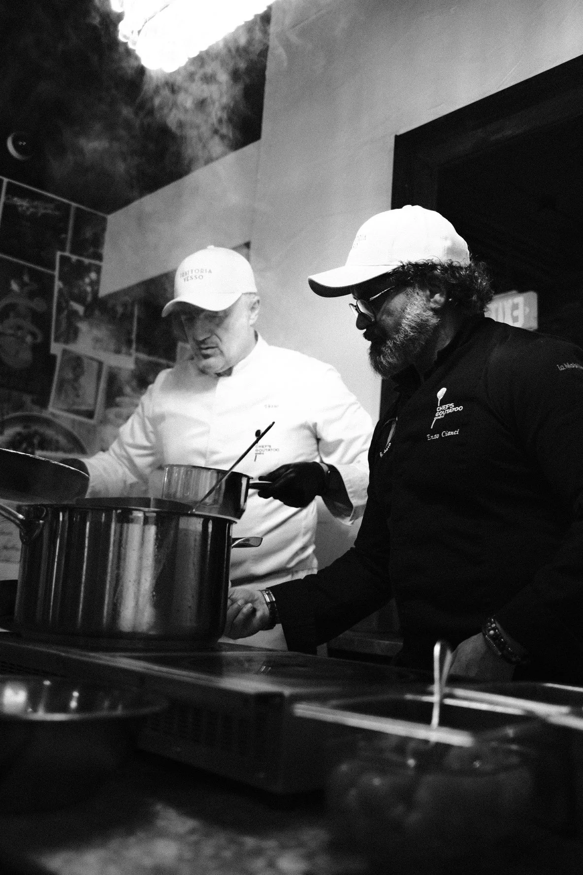 Two chefs in a professional kitchen, one wearing a white chef's coat and the other a black chef's coat, both with white caps, preparing food over a large pot, steam rising from it.