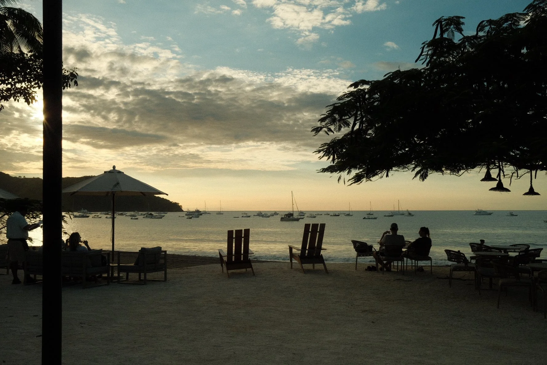 People relaxing on chairs and tables by a beach, with boats on the water and a sunset sky with clouds in the background.