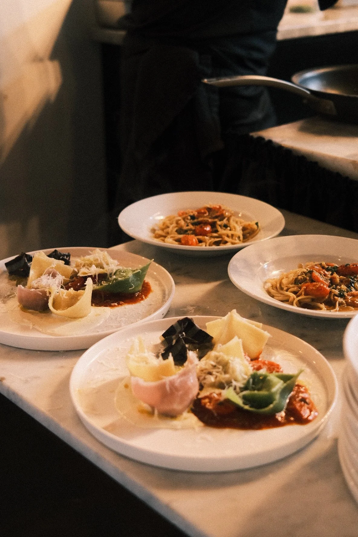 Plates of assorted Italian dishes, including pasta with tomato and vegetable sauce, and an antipasto platter with cheese, cured meats, and vegetables on a marble countertop.