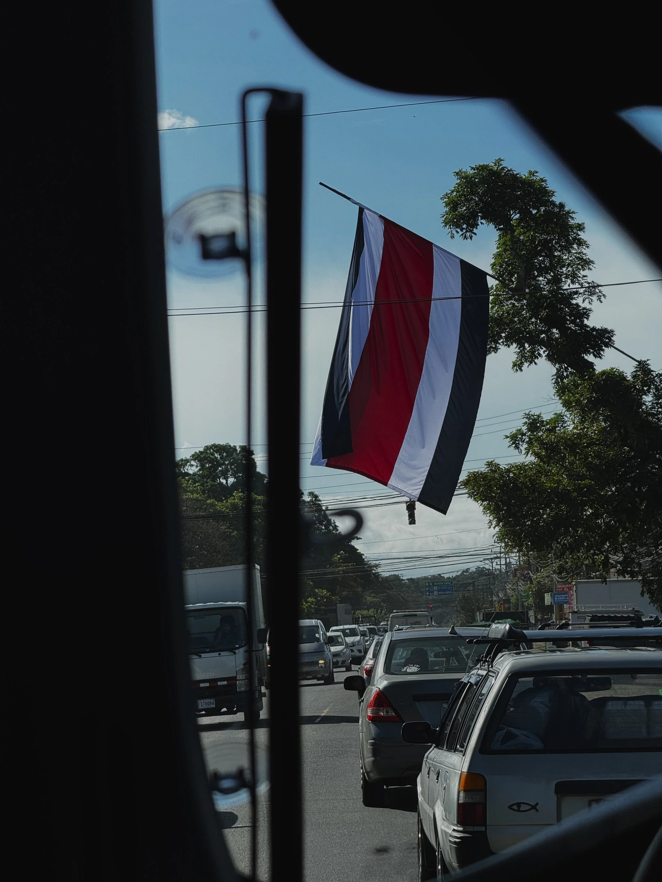 A busy street scene viewed through a vehicle window, with a large red, white, and black flag hanging from a pole against a blue sky and some trees.