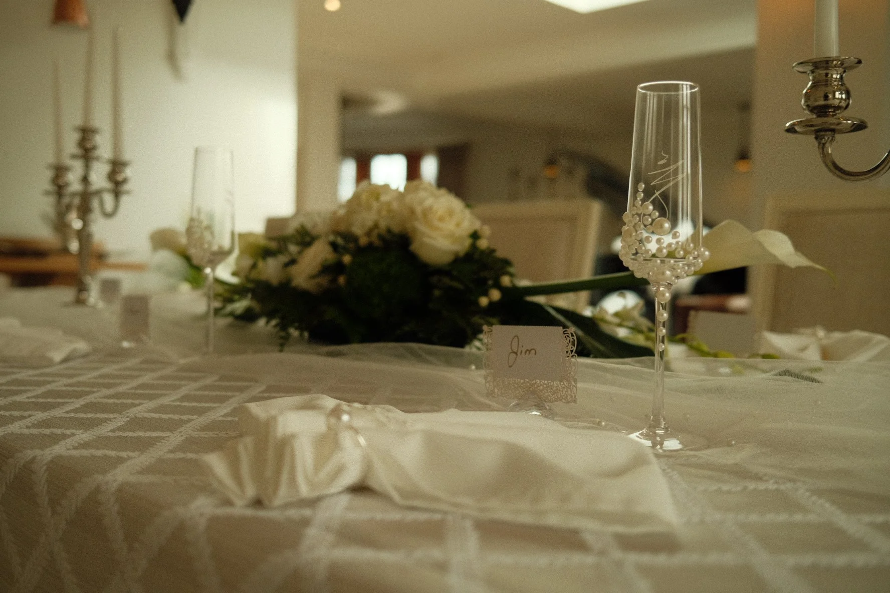 A decorated wedding table with champagne flutes filled with white decorative beads, a floral centerpiece with white roses and greenery, a small name card labeled 'Jim,' candle holders, and a cream-colored tablecloth.