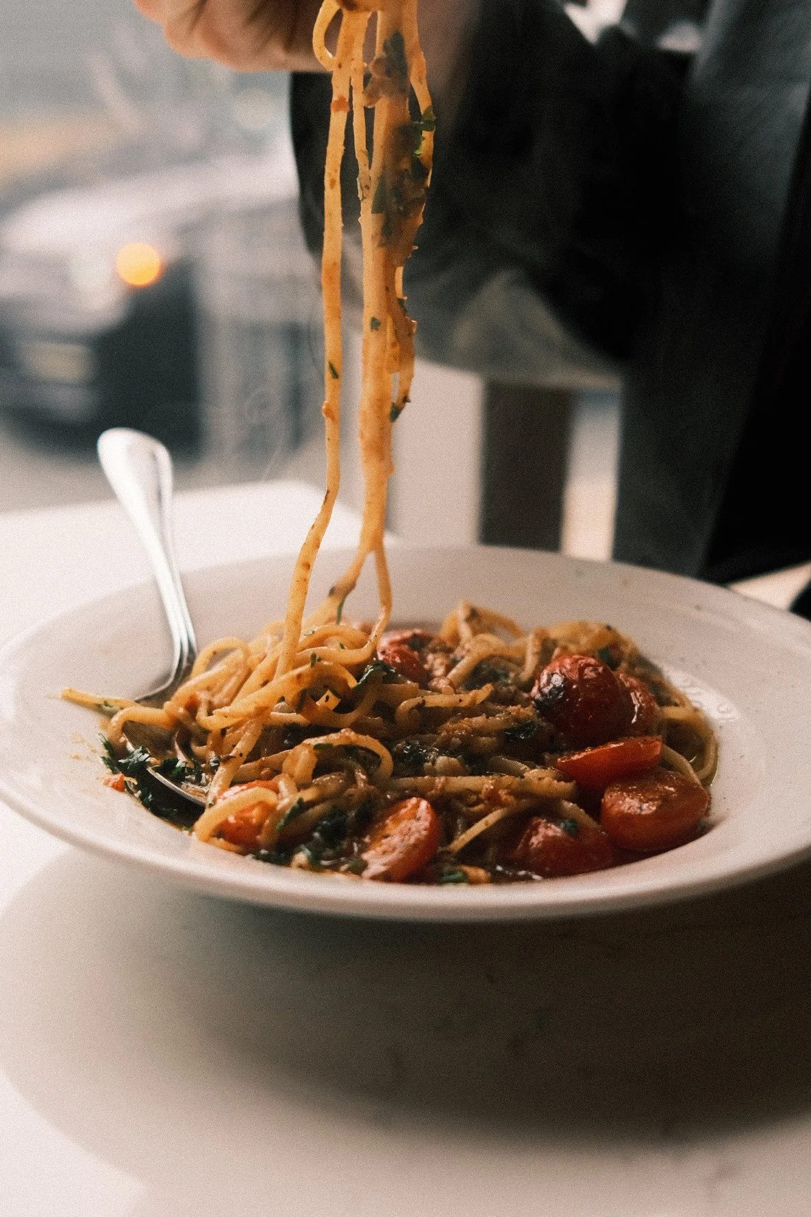 A plate of spaghetti with cherry tomatoes and herbs, with a fork resting on the side of the plate, and a person twirling some noodles with chopsticks.