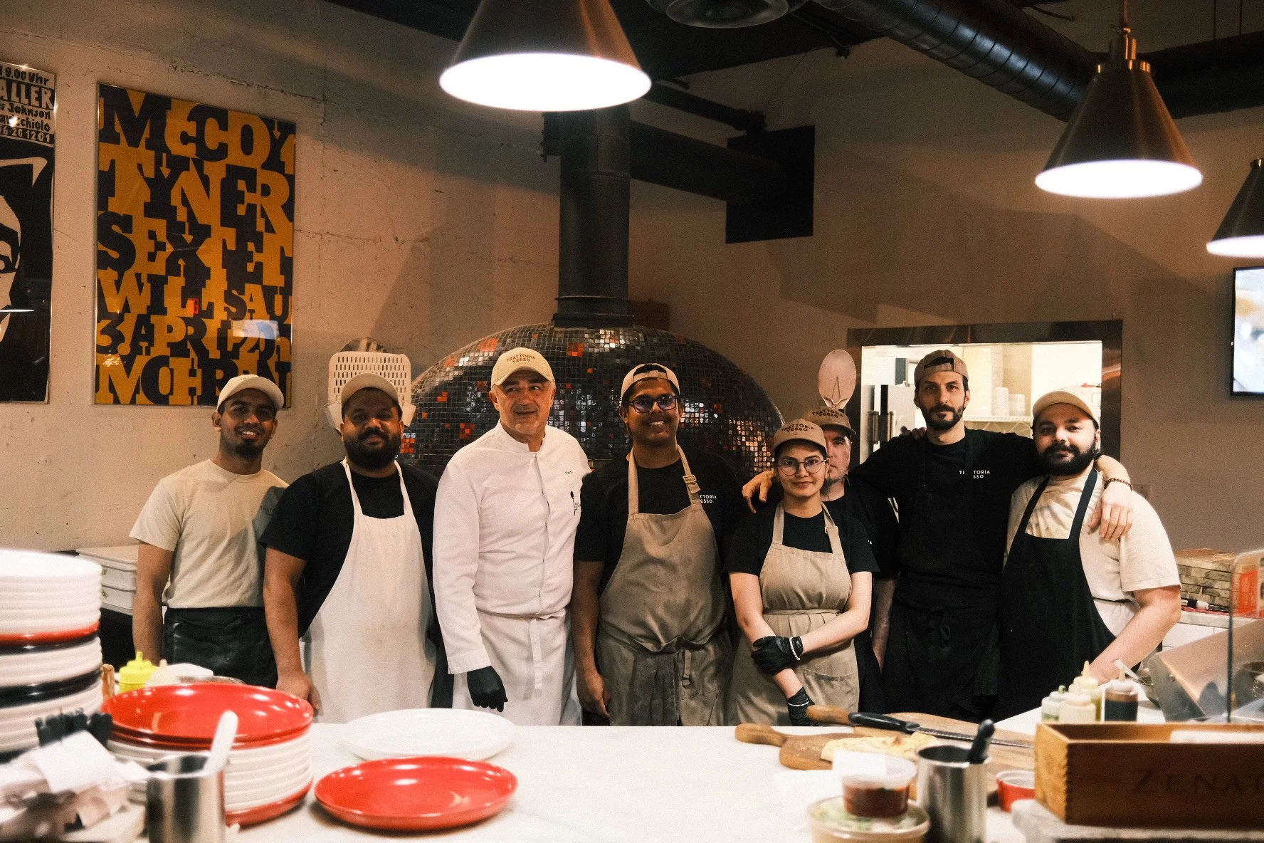 Group of nine diverse restaurant staff members, including chefs and kitchen staff, standing together in a restaurant kitchen with a large pizza oven in the background.