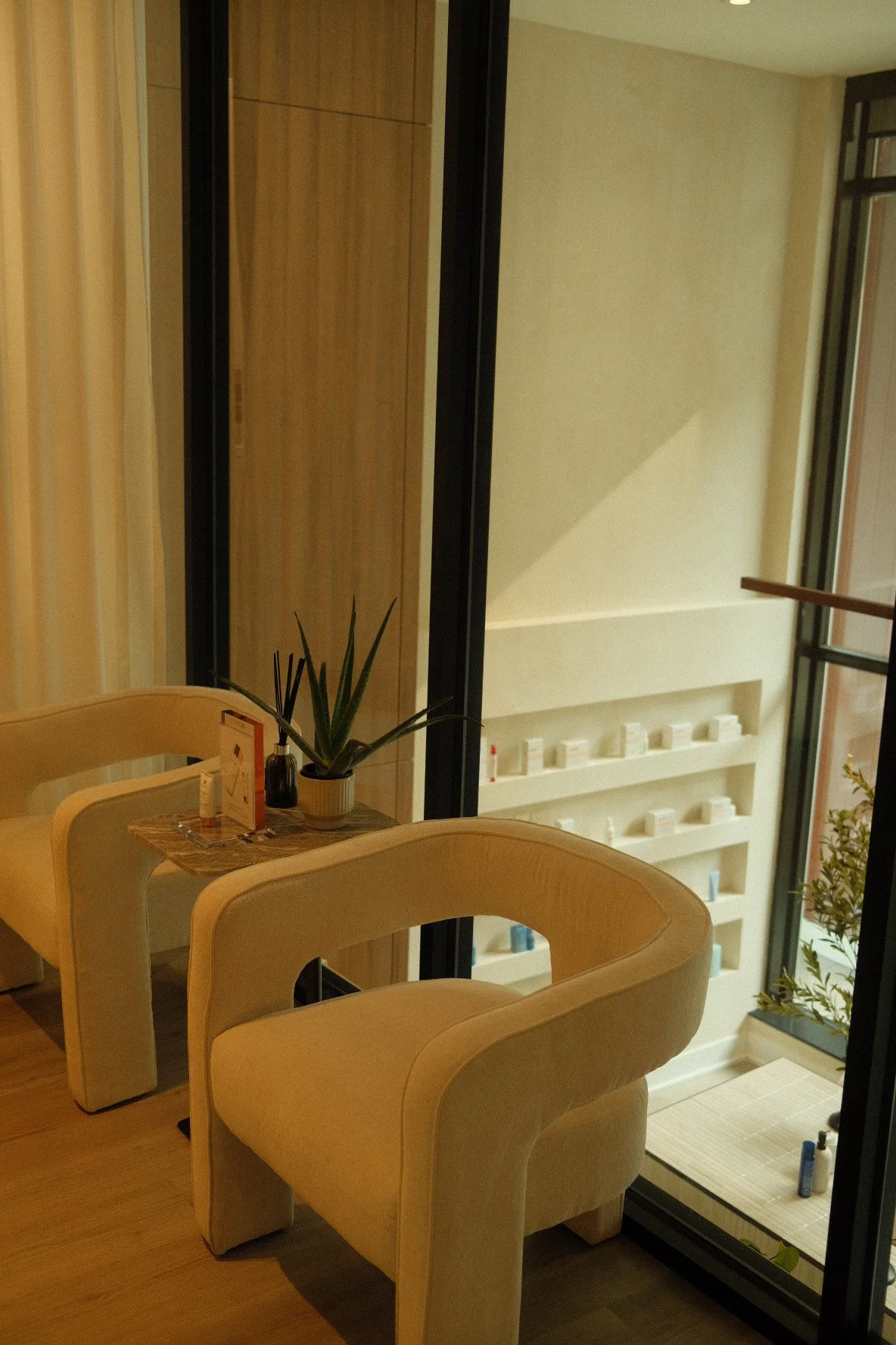 A cozy indoor seating area with two cream-colored armchairs, a small wooden table with a potted plant, and a bottle of lotion or sanitizer. Behind the chairs, a large sliding glass door shows a balcony with a white shelving unit containing small cont