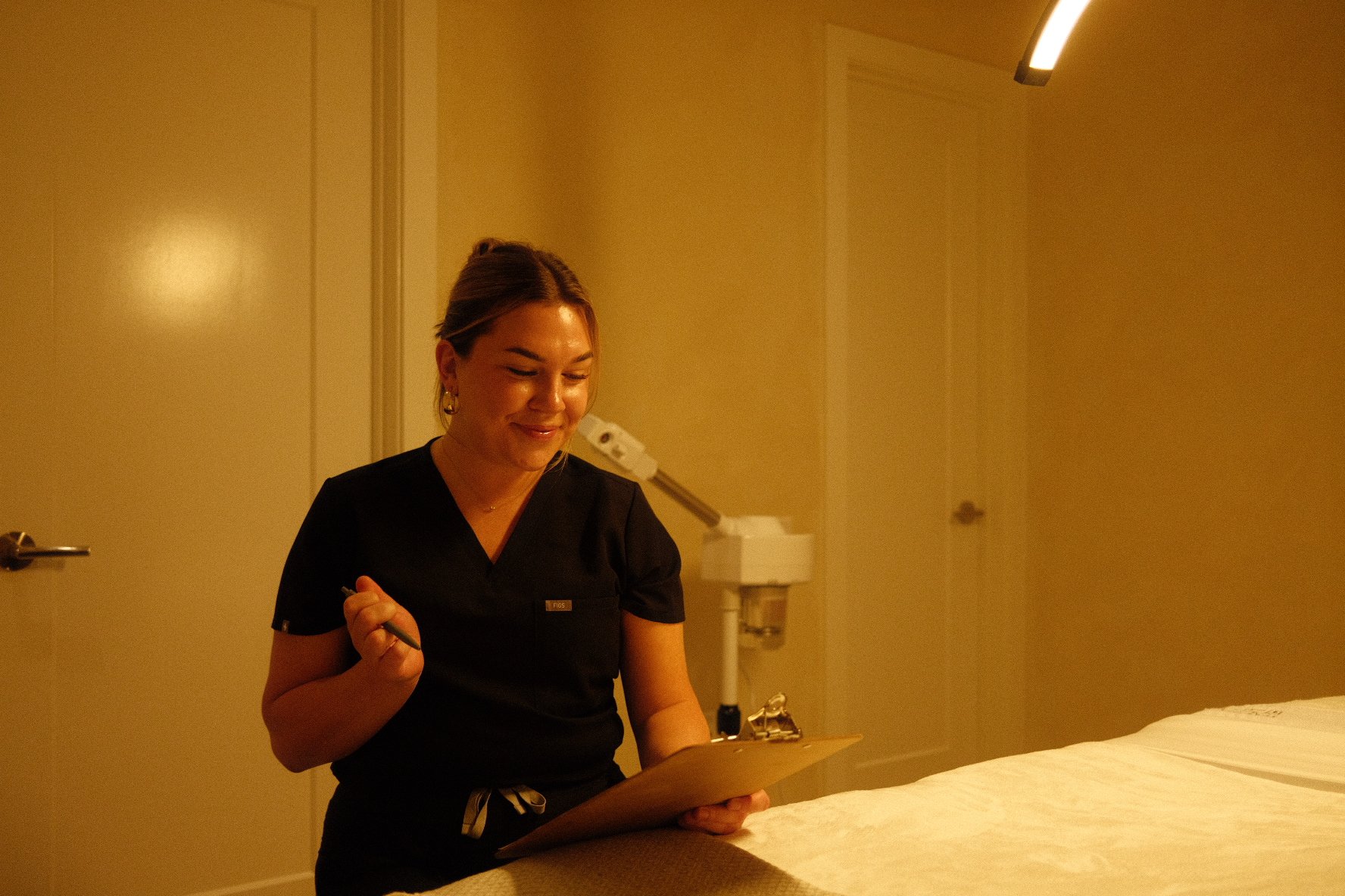 A woman in black scrubs smiling and holding a clipboard in a clinical or spa setting, with a beige wall and a door in the background.