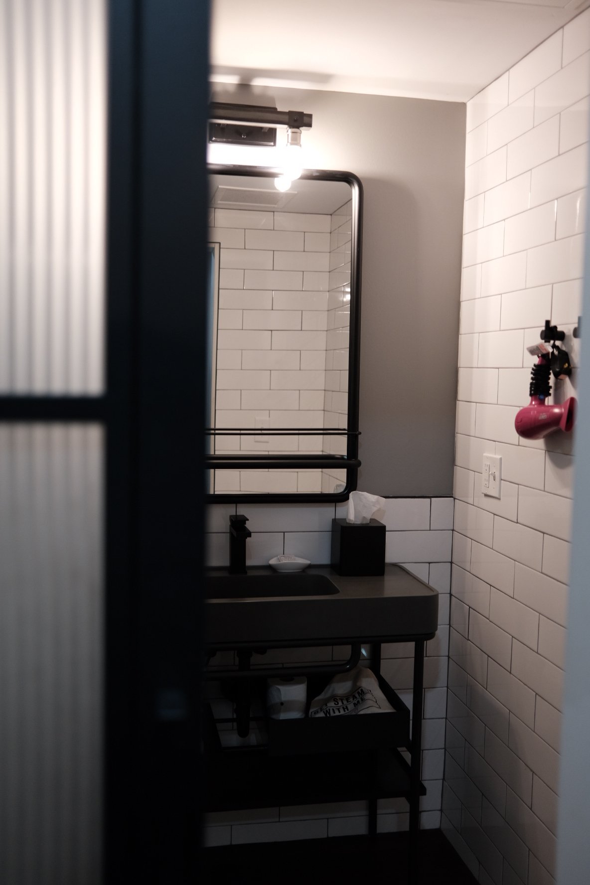 Small modern bathroom with white subway tiles, a black framed mirror, and black fixtures, including a small sink and shelving unit, with a tissue box and hand soap on the sink.