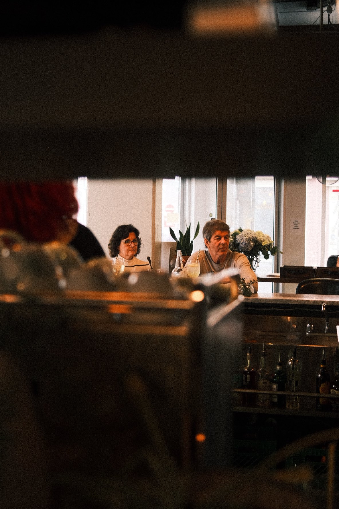 People dining inside a restaurant seen through a gap or opening, with one woman and one man visible, sitting at a table near large windows, with plants and bottles on the table, and other blurred figures or objects in the foreground.