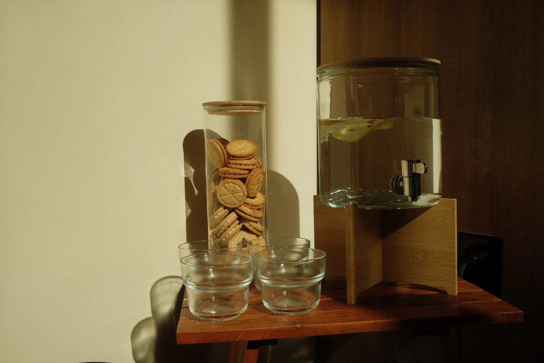 A wooden table with three empty glass cups, a glass jar filled with cookies, a large water container with lemon slices, and a wooden water dispenser.