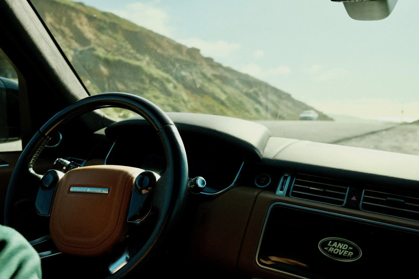 Inside a Range Rover vehicle, showing the dashboard, steering wheel, and a view of a winding road along a hillside from the front windshield.