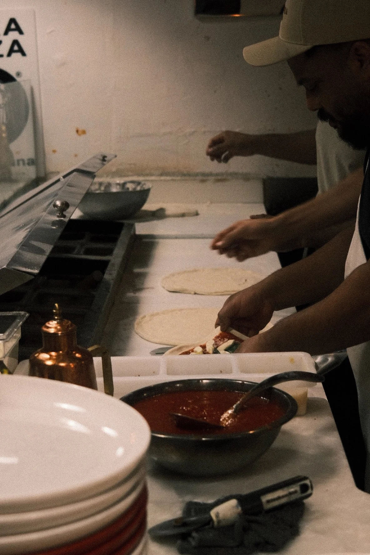 People preparing pizzas with toppings at a pizza restaurant kitchen counter. There are pizza doughs, a bowl of tomato sauce, and utensils visible on the counter.
