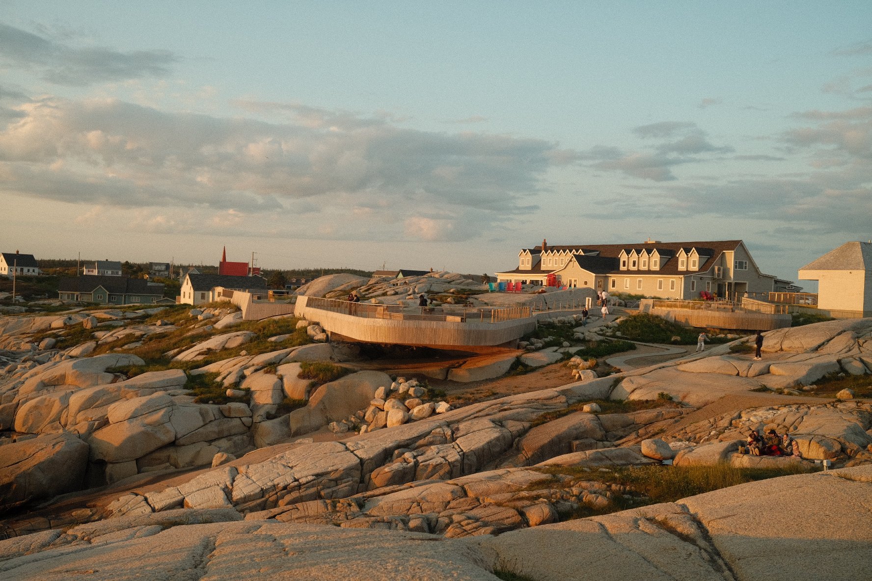 Rocky seaside with modern houses and a viewing deck, people walking and sitting on the rocks during sunset.