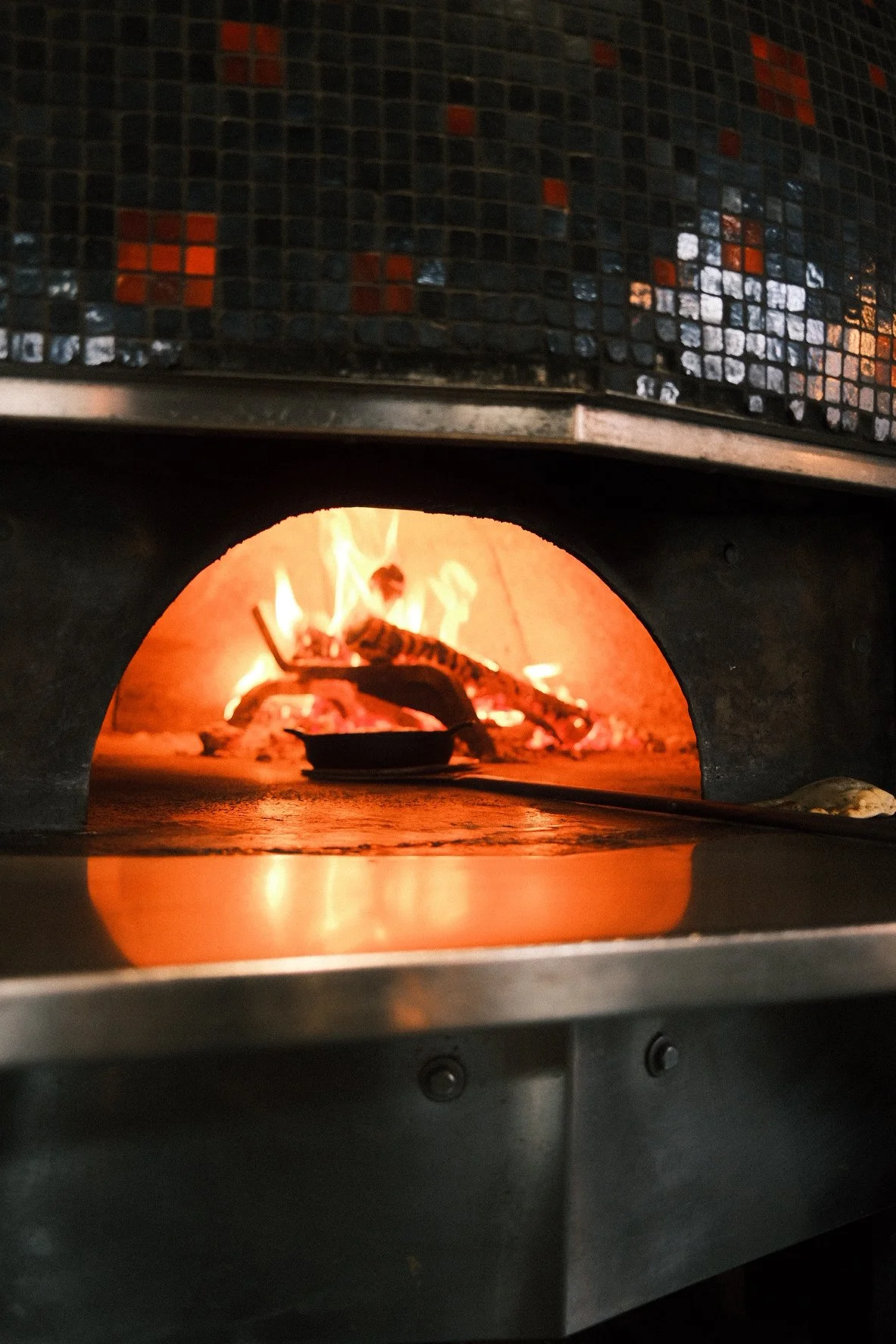 A pizza oven with a flaming fire inside, cooking a pizza on a black pizza peel.