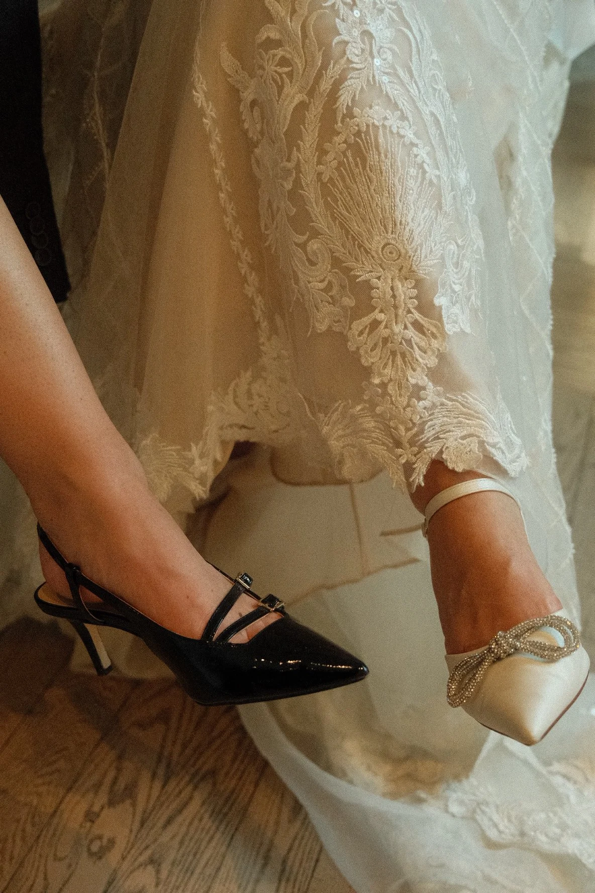 Close-up of a person's legs and feet, wearing a lace dress with intricate embroidery, one foot in a black high heel, the other in a white satin shoe with a decorative bow, on a wooden floor.