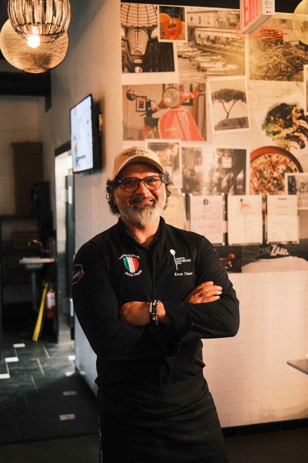 A man with glasses, a beard, and a mustache, smiling with arms crossed, wearing a black chef's jacket with Italian patches, a bracelet, and a beige cap, standing inside a restaurant with decorated walls and hanging lights.