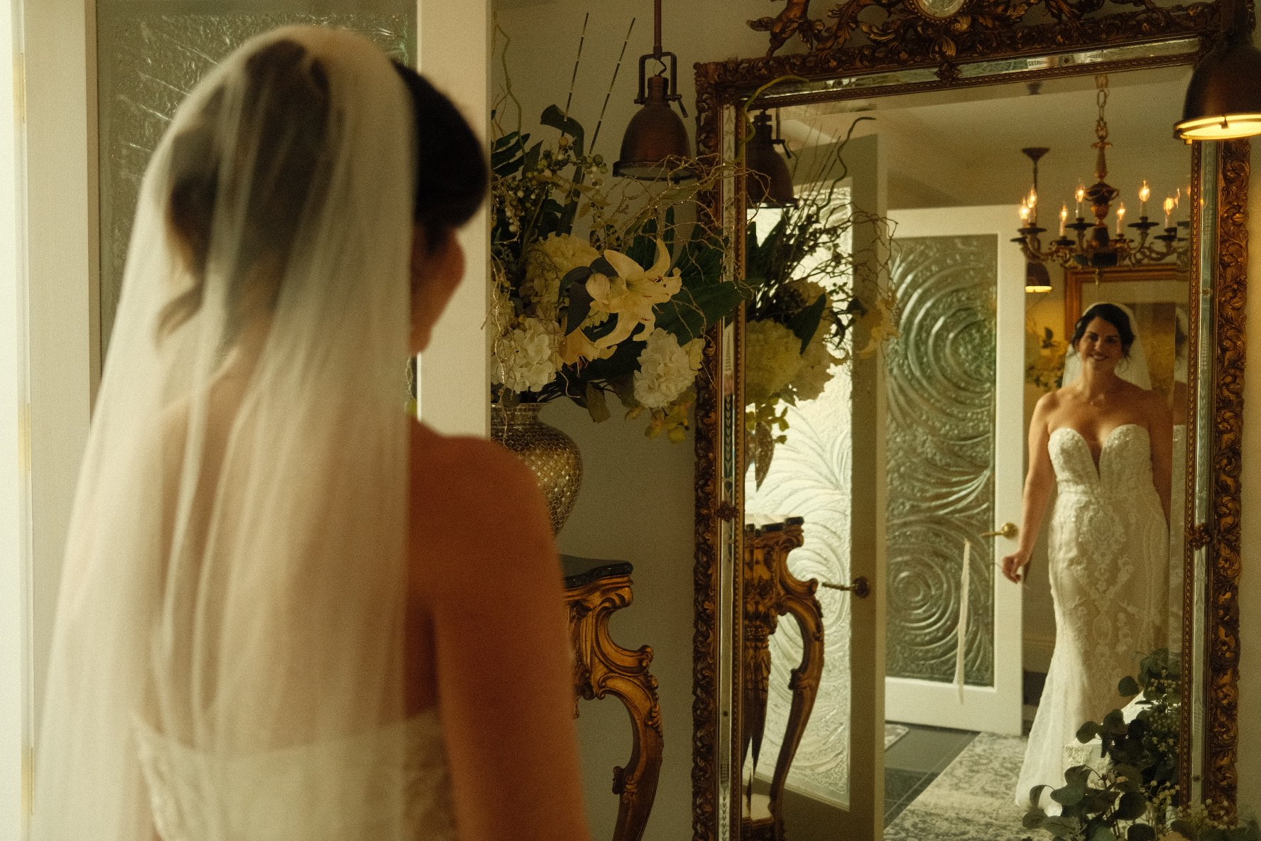 A bride in a strapless lace wedding gown looking at herself in a mirror, with her reflection visible, standing in an ornately decorated room with flowers and elegant furnishings.