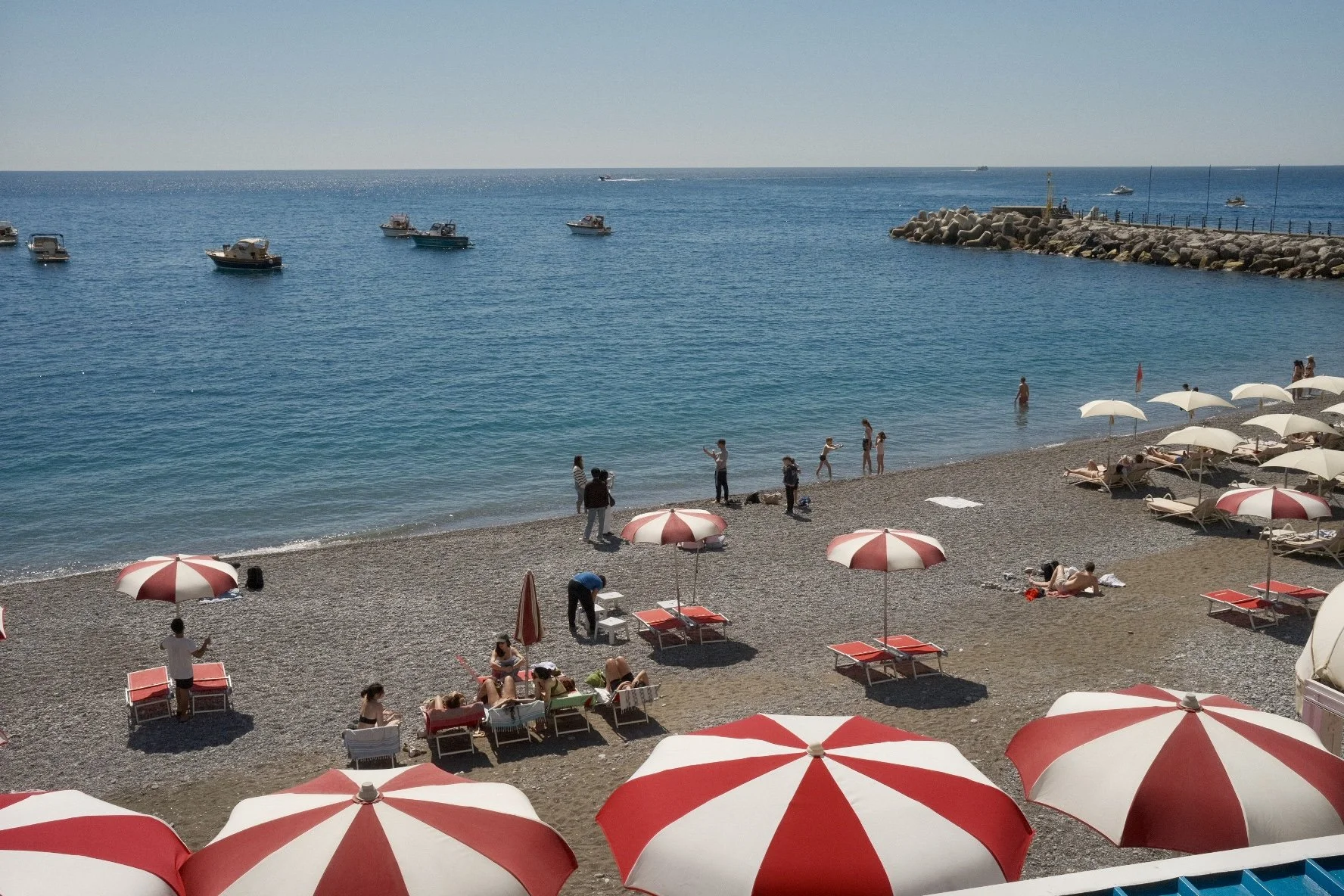 Beach scene with red and white umbrellas, sunbathers on lounge chairs, and people wading in the water. Several boats are anchored offshore, and there are people walking or standing along the shoreline with a breakwater and rocky pier in the backgroun