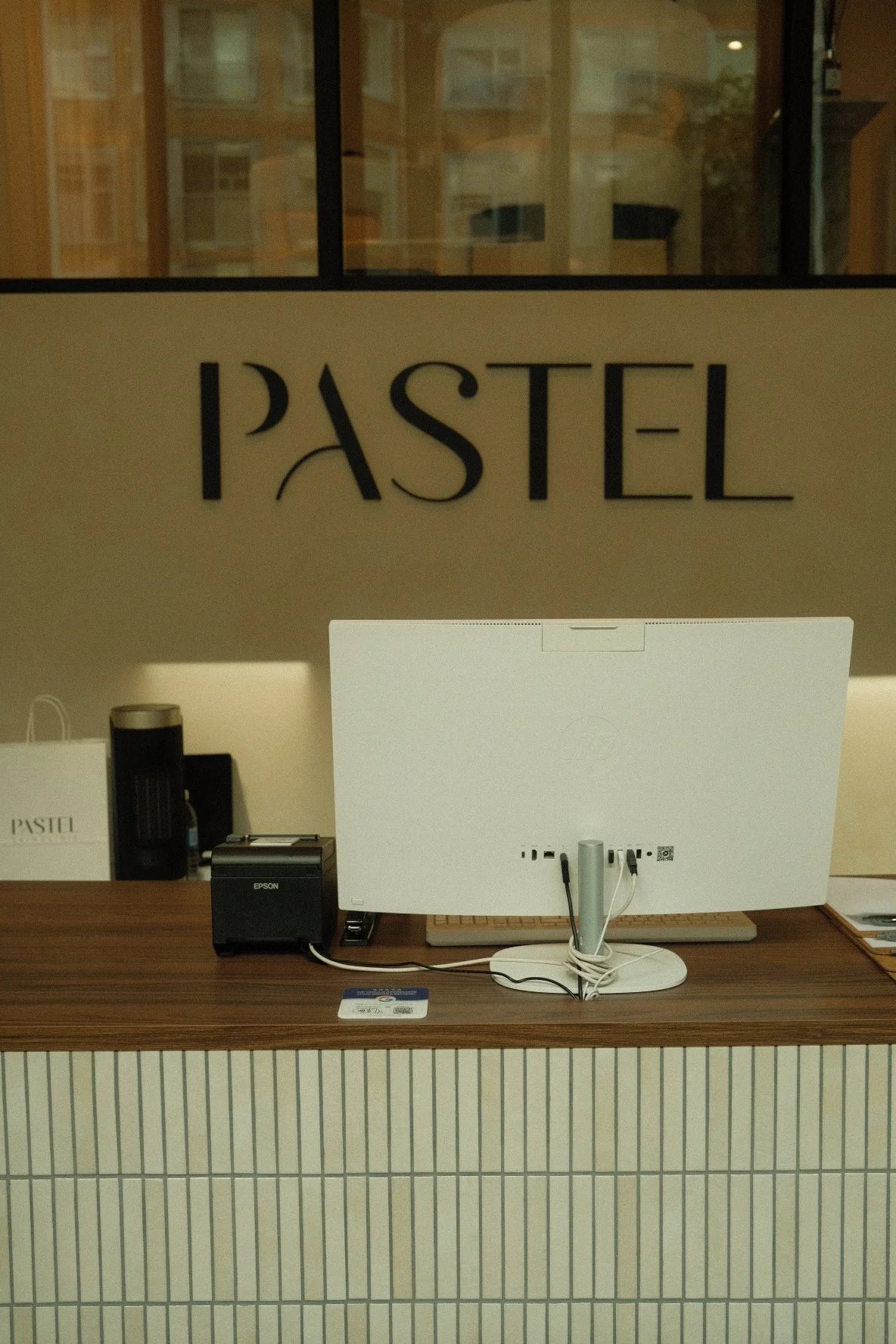 Hotel reception desk with a white computer monitor, inkjet printer, and a few small items, with a beige wall behind it displaying the word "PASTEL" in large black letters.