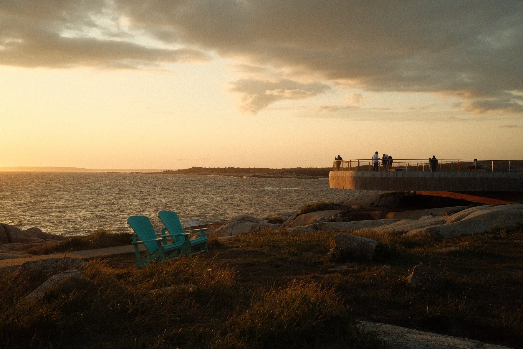 A coastal scene during sunset with surfers watching the ocean from a pier, two teal chairs on rocky ground, and a cloudy sky.