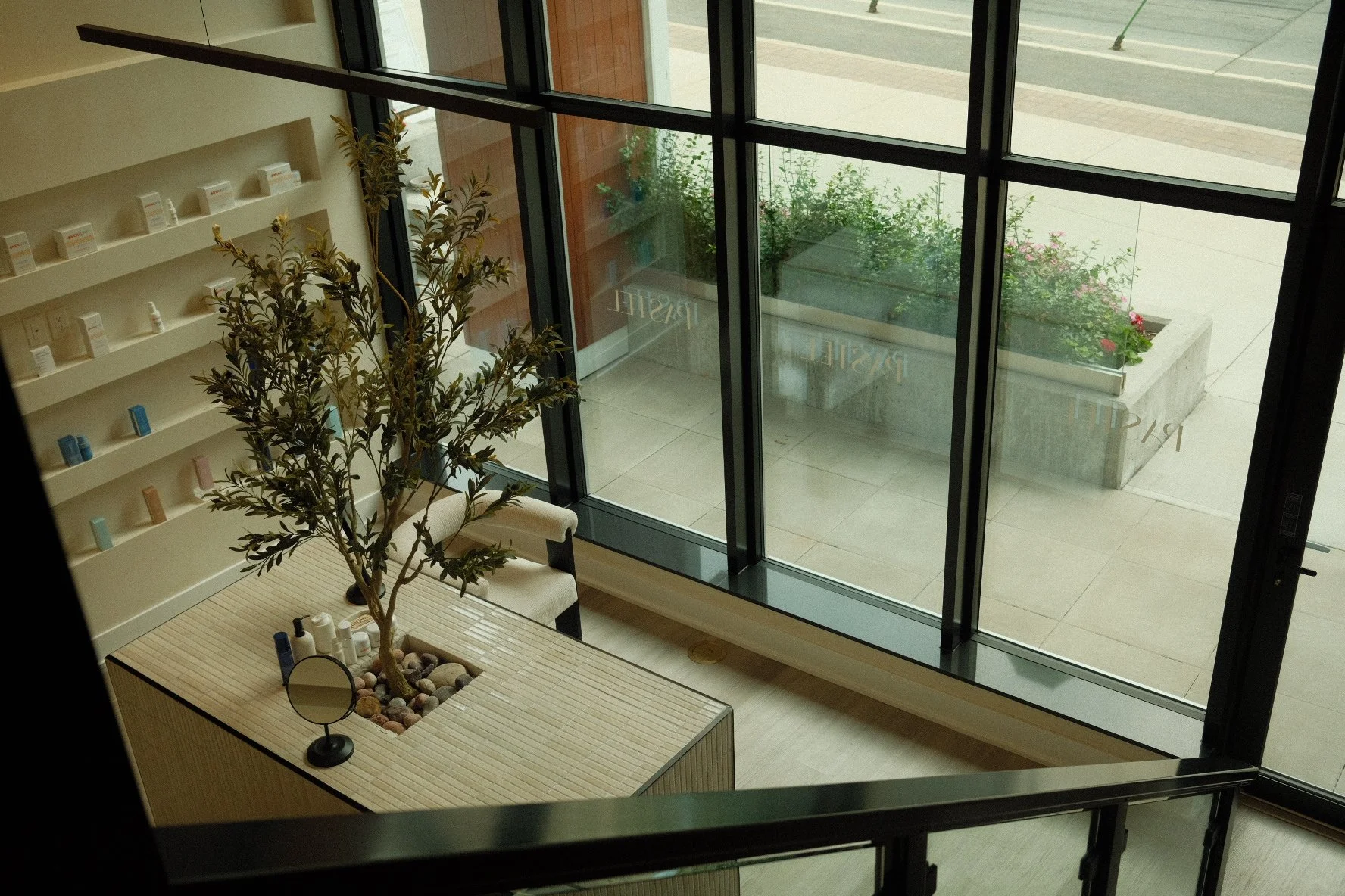 Interior of a modern building with large glass windows, a potted plant, and a table with skincare or cosmetic products.