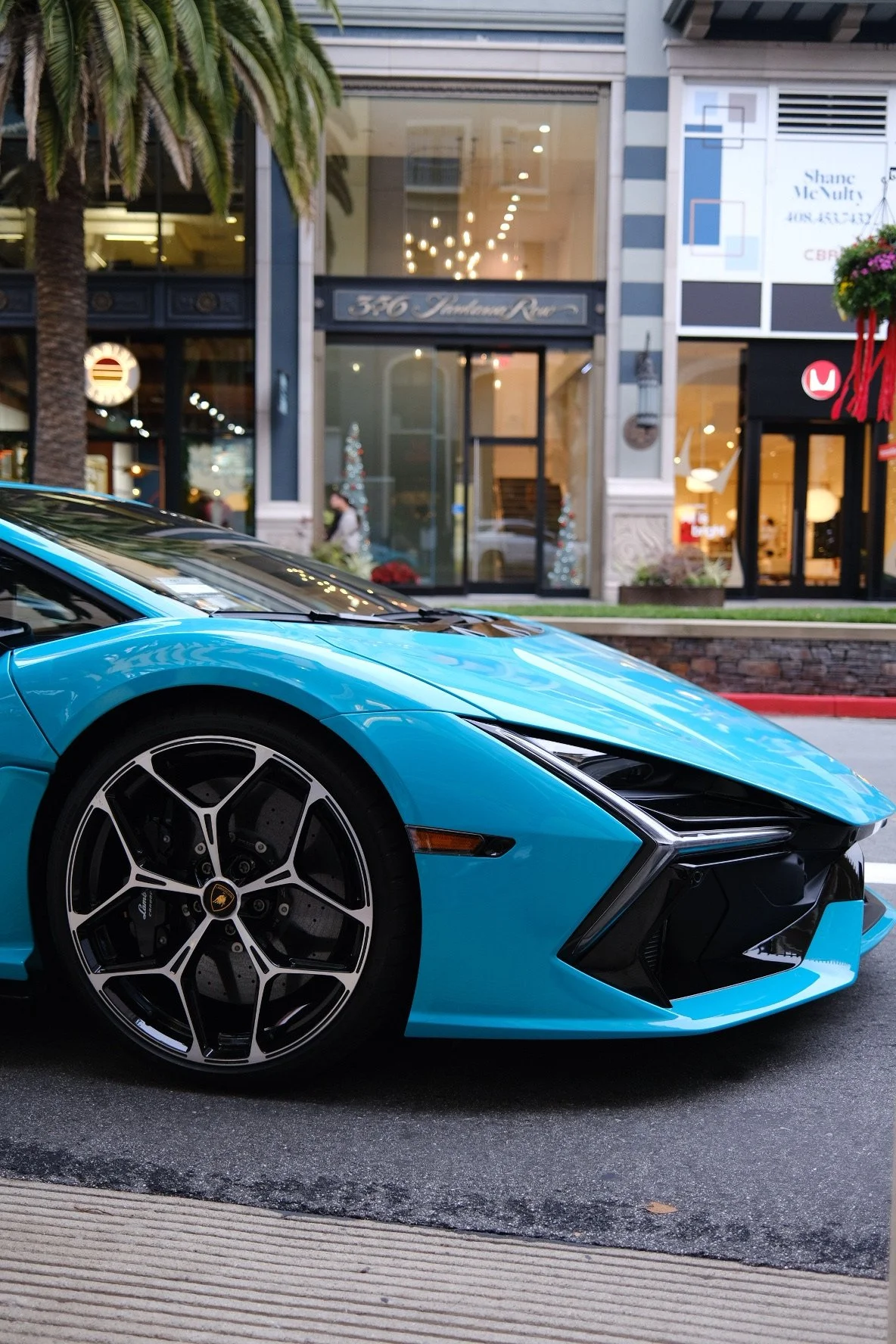 The image shows a blue Lamborghini supercar parked on the street in front of a shopping or office complex. The car is focus of the image, with the front part and wheel clearly visible, including the Lamborghini emblem on the wheel.