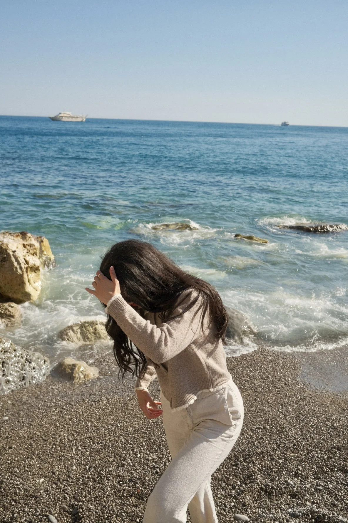 A woman with long dark hair, wearing a beige sweater and white pants, is standing on a pebble beach near the ocean, covering her face with her hand. The ocean has small waves, and there are two large boats in the distance against a clear blue sky.