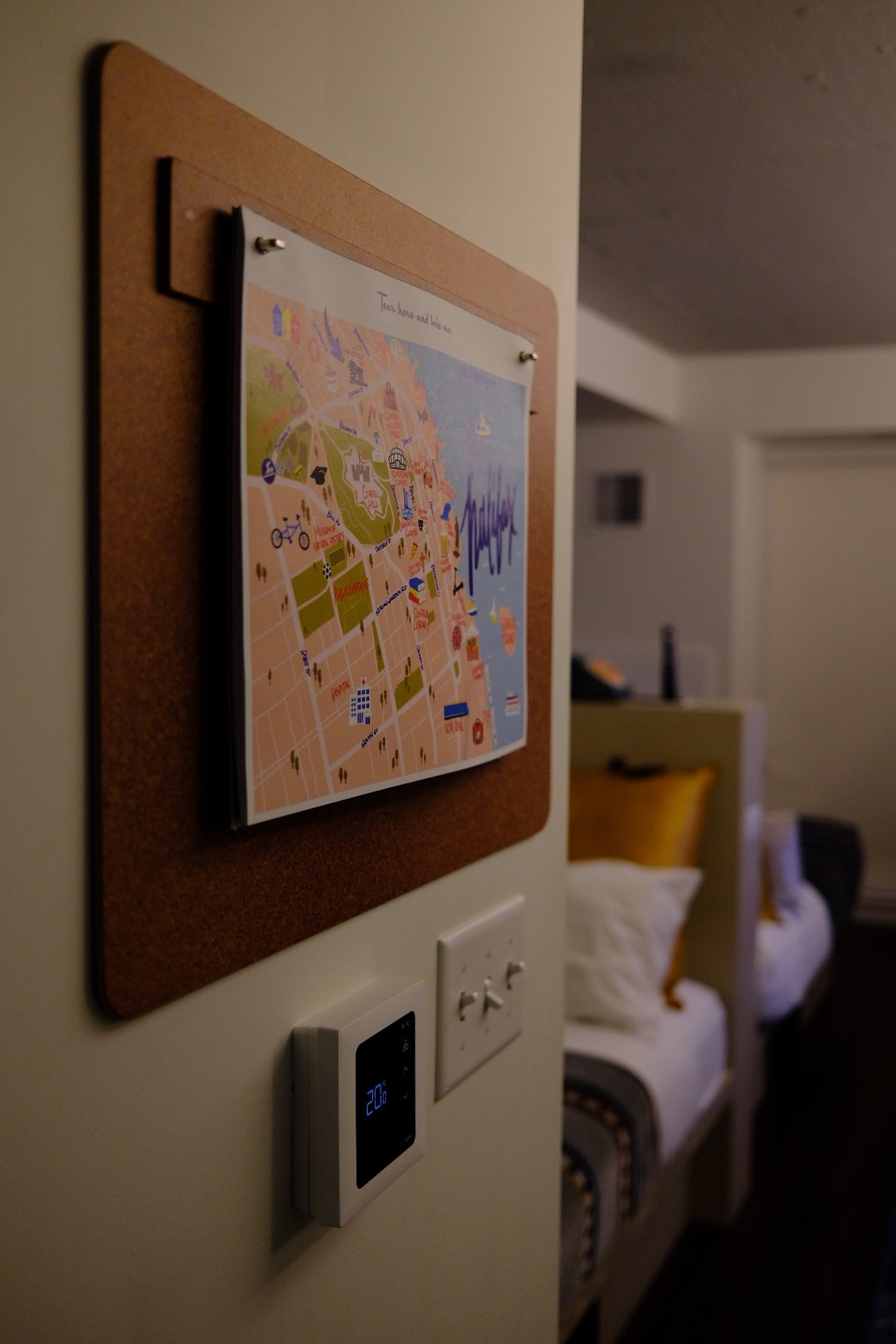 A cork bulletin board with a colorful map pinned to it, mounted on a wall with electrical outlets and switches below. In the background, a bed with a yellow pillow and a bedside table are visible in a dimly lit room.