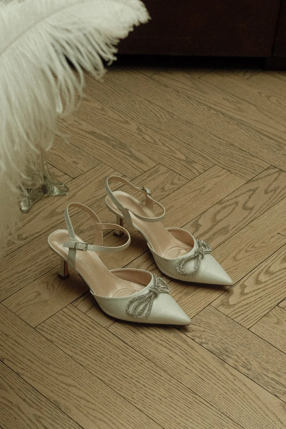A pair of elegant white high-heeled shoes with pointed toes, ankle straps, and decorative bows with pearls on the front, placed on a wooden floor.