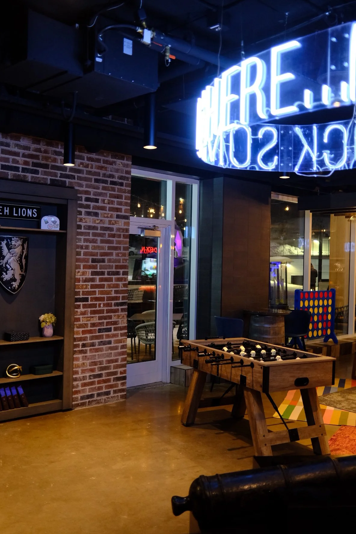 Interior of a modern bar or lounge area featuring a foosball table, brick walls, a neon sign, and a seating area visible through a glass door.