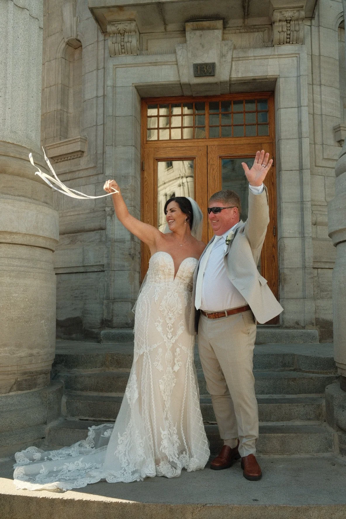 A bride and groom celebrating outdoors on steps in front of a large stone building with wooden doors, the bride in a strapless lace wedding gown with a veil and the groom in a beige suit, white shirt, brown shoes, and wearing sunglasses, both smiling