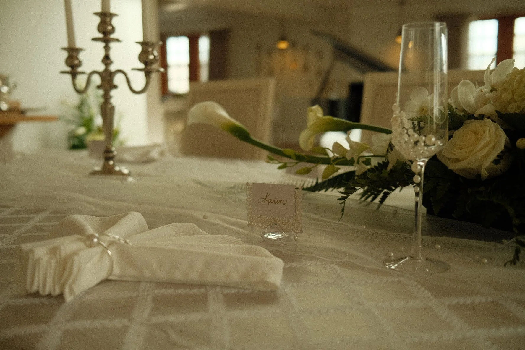 Elegant table setting with white flowers, a candelabra, champagne flute with pearls, and a place card that reads "Karyn" in a warmly lit room.
