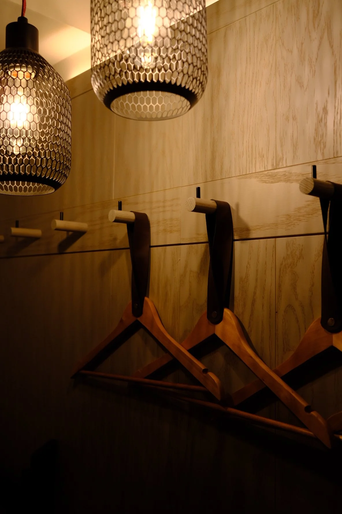 Wooden coat hooks and hangers on a wall illuminated by hanging pendant lights with a honeycomb pattern.
