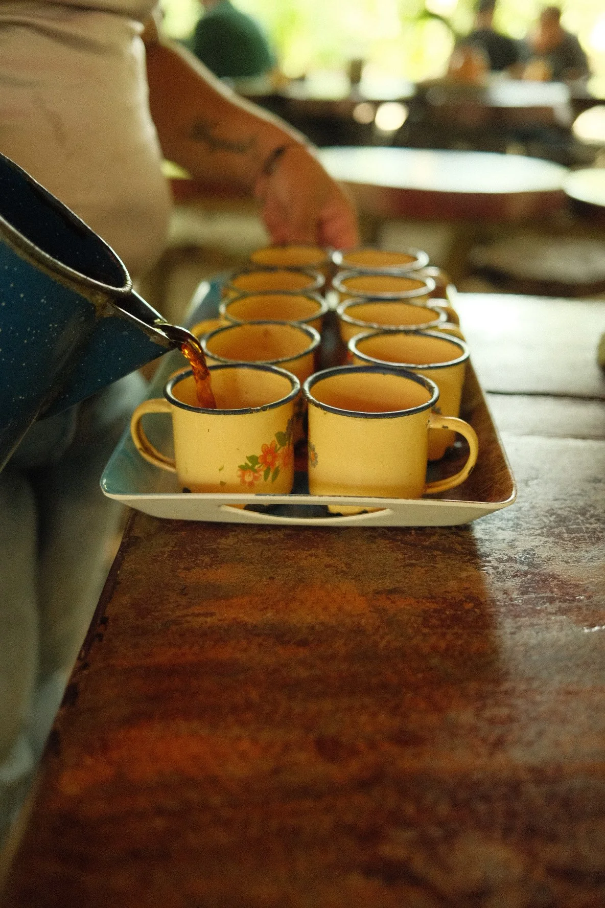 A person pouring a dark liquid, possibly coffee, into yellow ceramic cups with floral patterns arranged on a rectangular tray at a wooden table.
