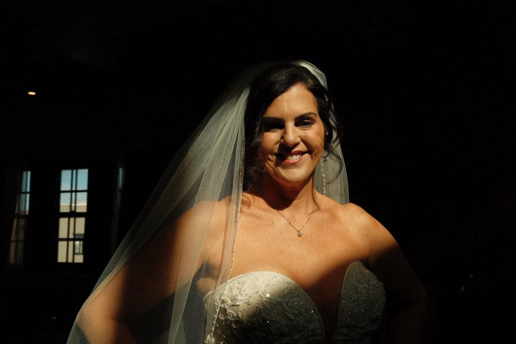 A woman in a wedding dress with a veil, smiling in a dark room with windows in the background.