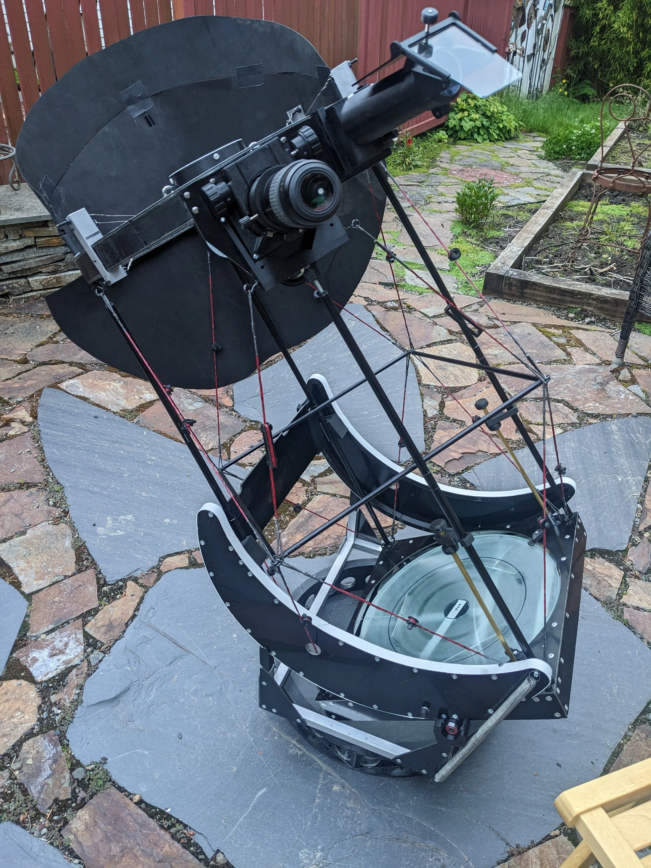 A large telescope with a black and white frame and red tension wires, positioned on a stone patio in a backyard, facing upward with the sky.