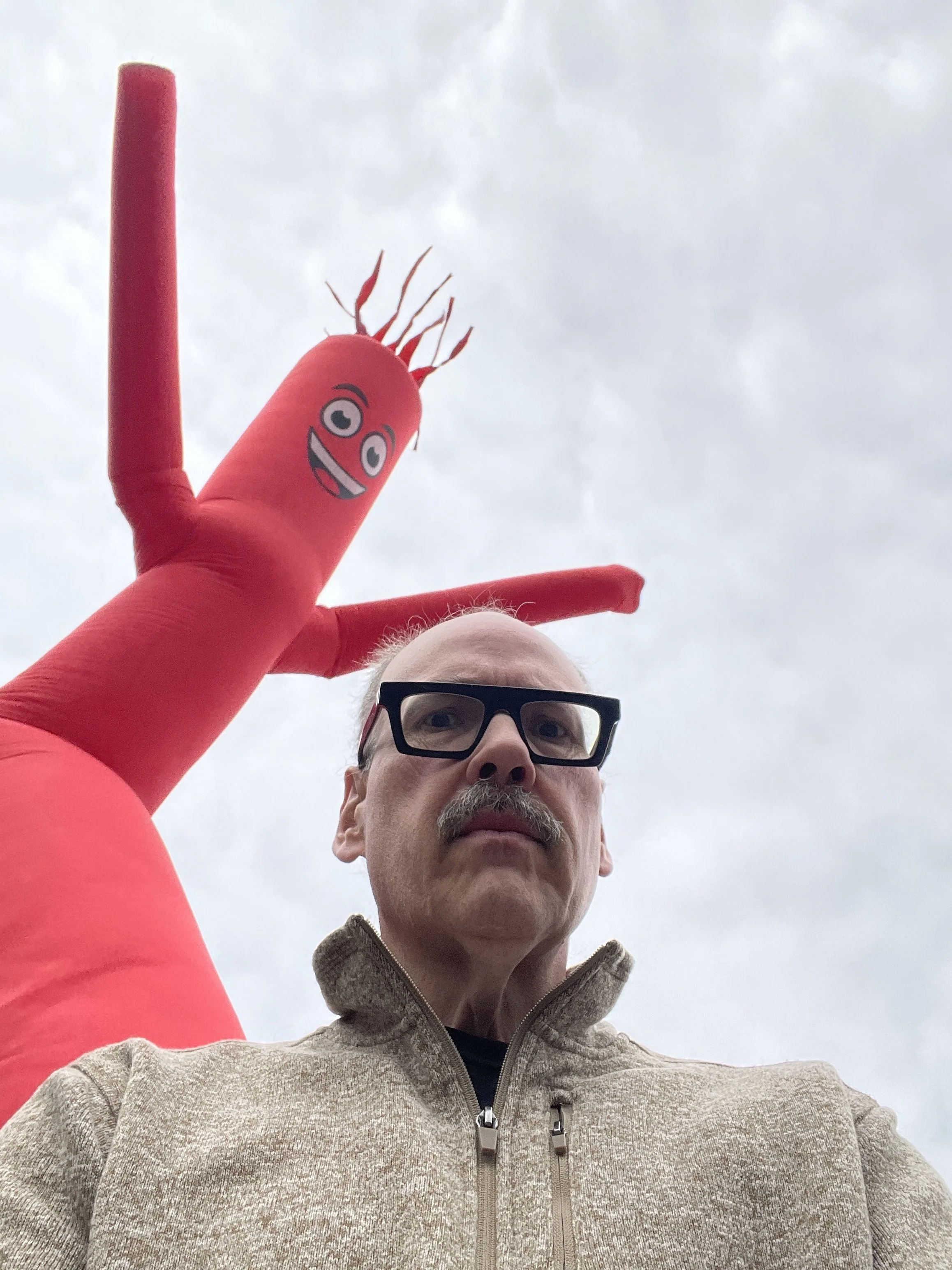 Wayne Rawley looking warily into camera with a large red inflatable tube man promotional balloon directly behind him.
