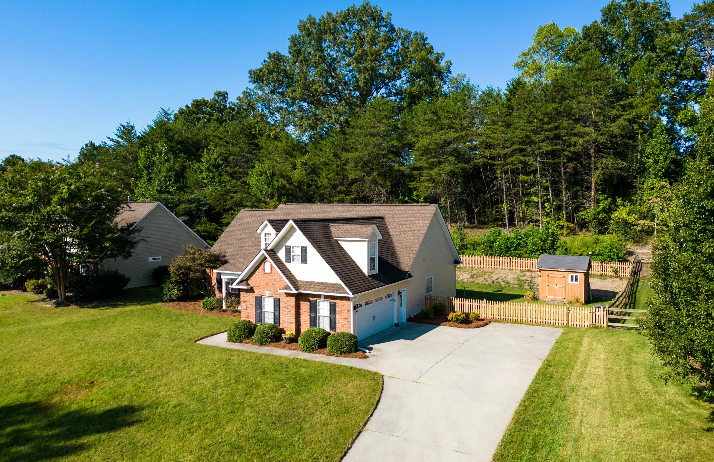 A beige two-story house with a brown roof, white garage door, and front porch. The house is surrounded by a green lawn, bushes, and a wooden fence. In the background, there are tall green trees and a clear blue sky.