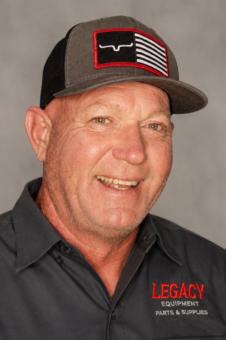 A smiling middle-aged man wearing a dark gray baseball cap with a Texas flag patch and a black work shirt with red embroidered text reading 'LEGACY EQUIPMENT PARTS & SUPPLIES'.