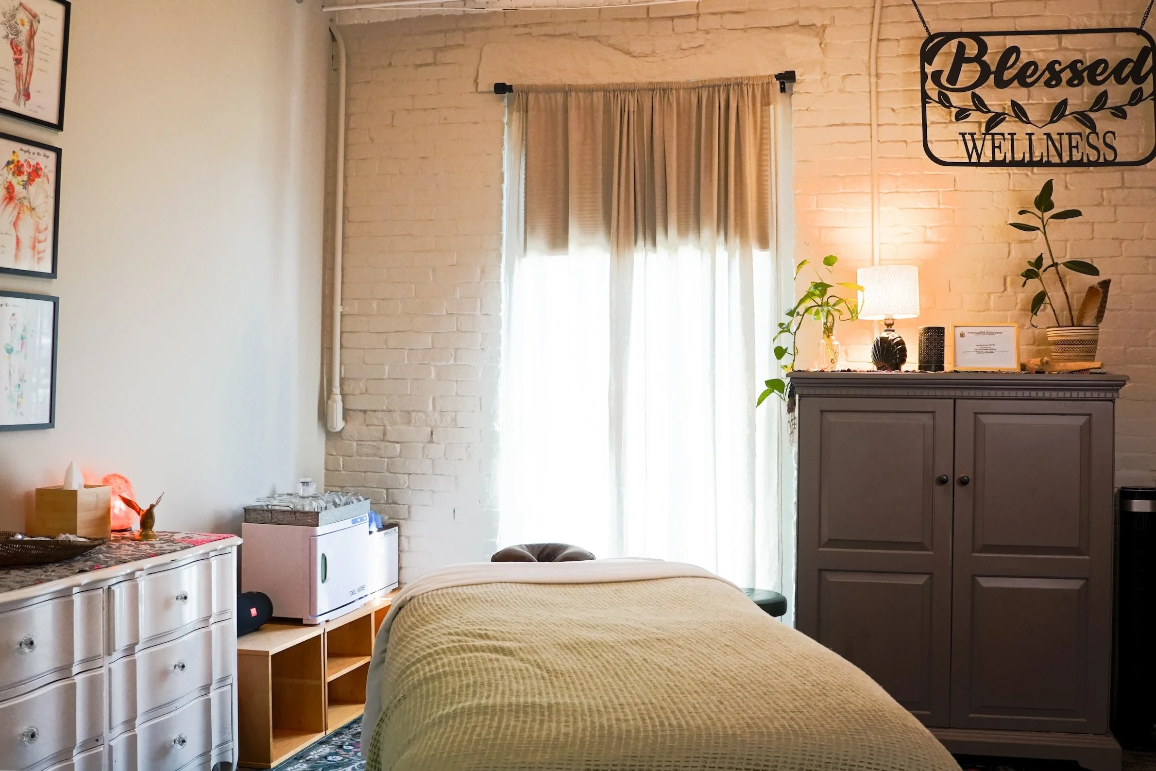 A cozy wellness room with a bed covered in a beige blanket, a white dresser with multiple drawers on the left, a small mini fridge underneath, and decorative artwork on the wall. A window with beige curtains allows natural light to brighten the room. A gray cabinet with plants, a lamp, and framed certificates are on the right wall. A sign reading 'Blessed Wellness' is hanging on the wall above the cabinet.