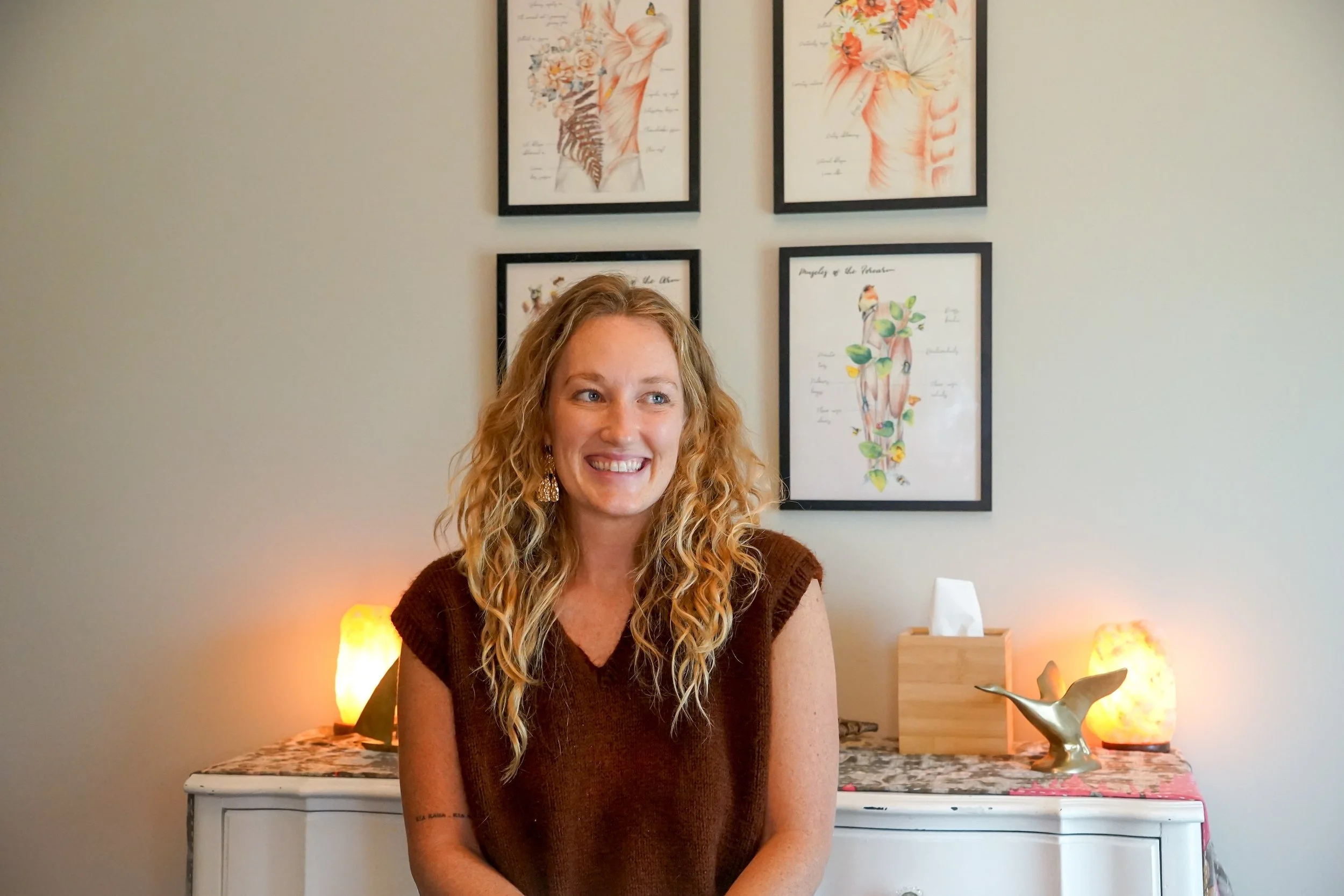 A woman with curly blonde hair and a brown top smiling and sitting in a room with a light-colored wall. Behind her, there are four framed art prints depicting anatomical and botanical illustrations, and the room is decorated with salt lamps and a wooden sculpture on a white dresser.